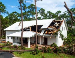Severe wind storm damage to a white house with a collapsed roof and fallen trees surrounding the property.